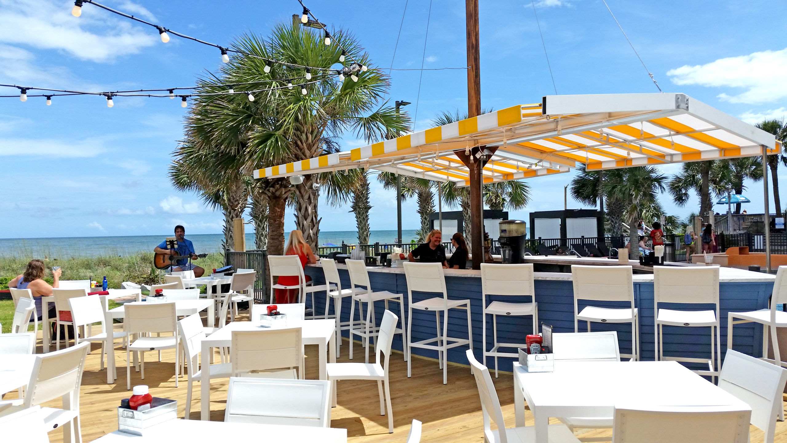 A beachside bar with white tables and chairs, a yellow and white striped awning, and palm trees in the background.